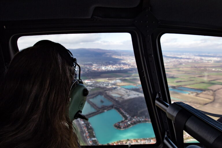 Blick aus einem Helikopter, Person mit Headset schaut auf eine Landschaft mit Seen und Feldern.