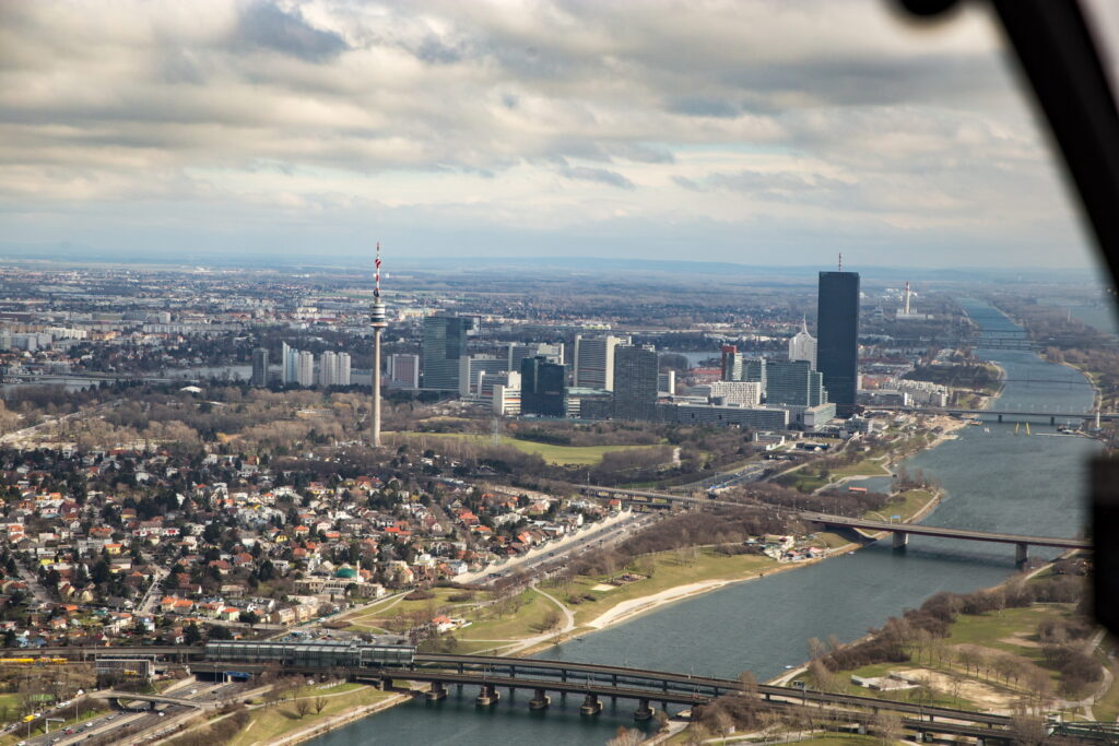 Wien aus der Luft mit Donauturm, modernen Hochhäusern und Donau, umgeben von Wohngebieten und Brücken. Im Hintergrund UNO City und Donauinsel.