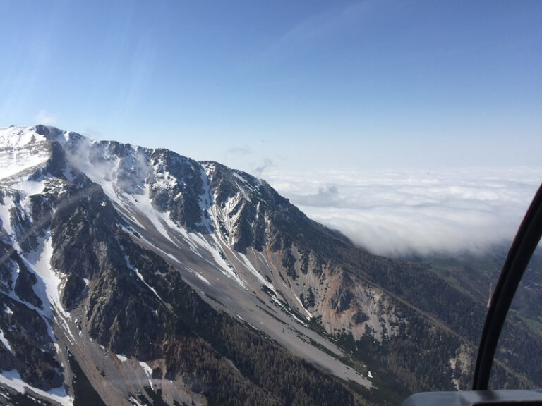 Berggipfel mit Schneefeldern und steilen Hängen, darüber ein klarer Himmel und tiefliegende Wolken.